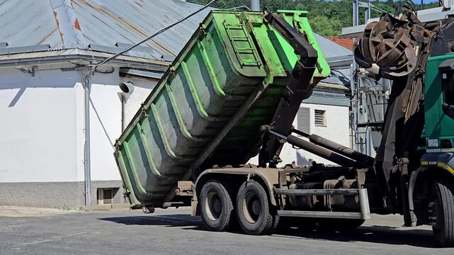 Heavy duty hooklift truck loading a large green metal waste container at an industrial site. Hydraulic mechanism operating roll-off dumpster for garbage removal, recycling, and construction waste mana