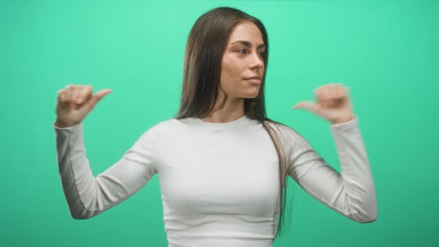 Woman pointing thumbs to self in studio against green screen wearing white long sleeve top and long hair; confidence.