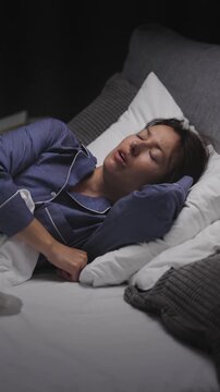 A woman in her thirties is lying in bed wearing blue pajamas while trying to sleep without success, tossing and turning under a dim light in a bedroom filled with pillows.
