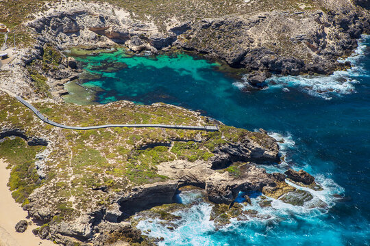 Aerial view of Fish Hook Bay with turquoise waters, rugged rocky coastline, and a wooden boardwalk winding through coastal vegetation, Rottnest Island, Western Australia, Australia.