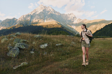 Fototapeta premium Young woman hiking outdoors in casual clothes with backpack walking on grassy trail near rocky mountain under clear sky, active lifestyle and nature exploration