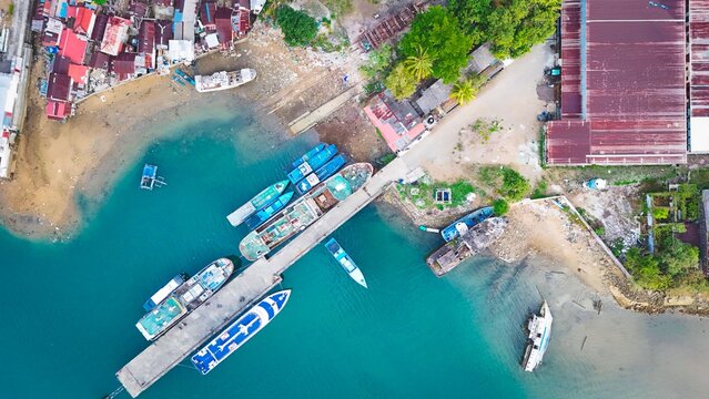 Aerial view of boats docked at a pier in a coastal area with turquoise water, surrounding buildings, and lush vegetation in Kecamatan Sirimau, Maluku, Indonesia.