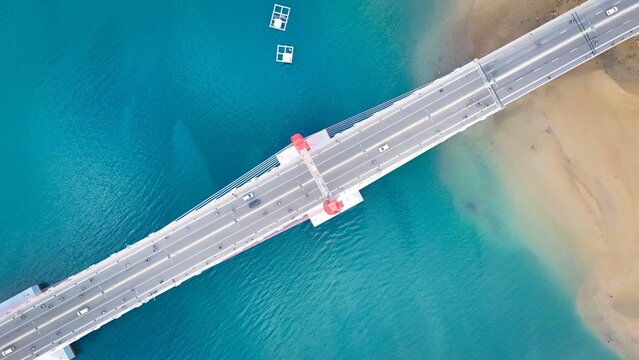 Aerial view of Jembatan Merah Putih spanning over turquoise waters with sandy shores and traffic in Kecamatan Sirimau, Maluku, Indonesia.