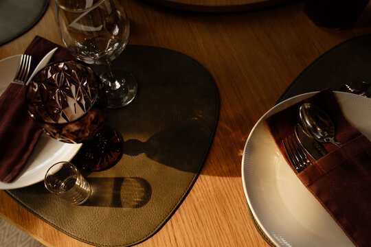 Warmly lit table arrangement featuring patterned goblet, clear wine glass, white textured plates, silver cutlery, burgundy napkin, leather placemats, and oak surface with refined dining style
