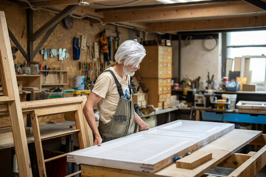 Skilled master puts antique wooden interior door on worktable in small business carpentry workshop, professional woodworker preparing vintage item for renovation, work preparation stages in studio.