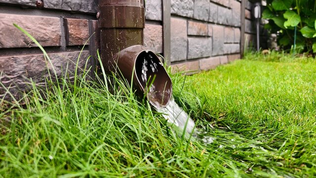 It is raining heavily, and water is draining from the roof of the house along the wall through a downpipe. The drops scatter across the grassy lawn. Cloudy summer weather