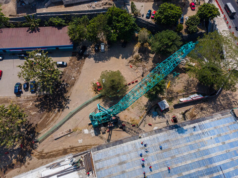 Aerial view of a construction site featuring a large teal crane, workers on a blue-tarped roof, and surrounding trees and vehicles in the Special Region of Yogyakarta, Indonesia.
