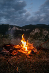 Glowing campfire in a mountain landscape at dusk beneath a dramatic cloudy sky. Perfect for themes of camping, outdoor adventure, hiking, travel, wilderness, survival, and peaceful evenings in nature. © VSzili