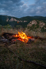 Glowing campfire in a mountain landscape at dusk beneath a dramatic cloudy sky. Perfect for themes of camping, outdoor adventure, hiking, travel, wilderness, survival, and peaceful evenings in nature. © VSzili