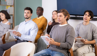 Fototapeta premium Middle aged man student at lecture listens to teacher and takes notes of data. Multinational group of students with notebooks and tablets during lesson at foreign language school, acting school. .