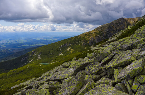 Epic alpine landscape with lichen-covered rocks in foreground, a tarn, and deep valley in the High Tatra Mountains.