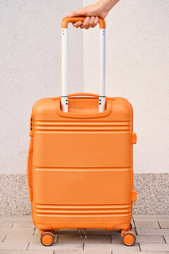 Orange rolling suitcase with extended handle held by person against light wall