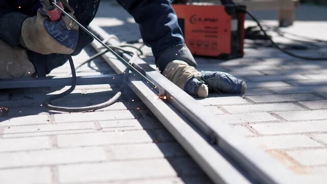 Outdoor worksite close view shows gloved worker welding along metal rail on paved surface, bright sparks at seam, cable in foreground, blurred orange equipment behind under hard daylight and cool