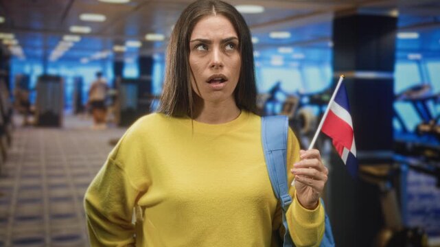 Woman holding dominican flag with right hand, open mouth, blue backpack strap visible while standing in studio gym area; surprise national pride.