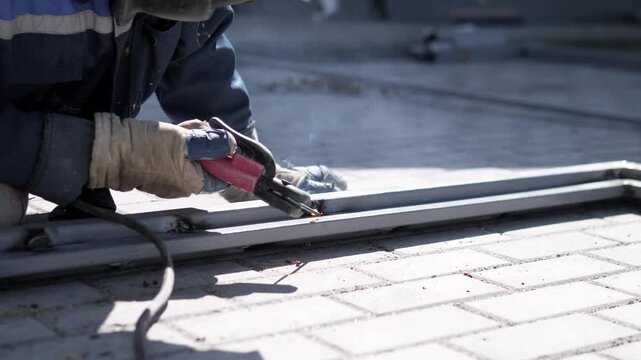 Close-up industrial scene shows protective gloves, welding tool, bright metal bars, and scattered sparks over paved ground, with smoke, hard light, shallow focus, and rugged worksite mood
