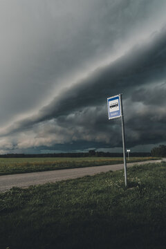 View of the Saxby bus stop sign standing beside a rural road under a dramatic, dark, stormy sky over green fields in Saxby, Laane County, Estonia.