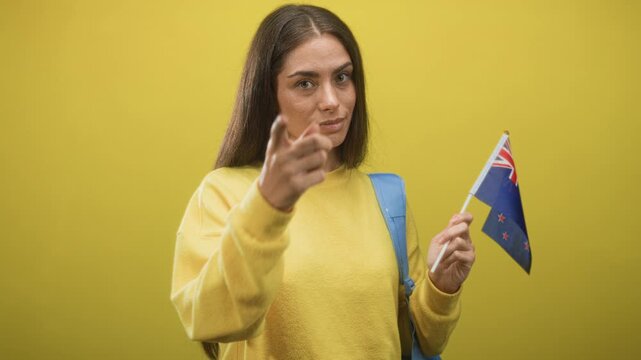 Woman wearing yellow sweater and backpack holding new zealand flag points finger at camera in studio; study abroad confidence.