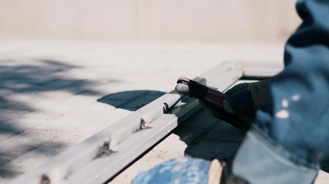 Close-up shows gloved hand holding pry bar against painted wooden trim during exterior repair work, with slight leverage at board gap, partial body at right edge, soft daylight, blurred background