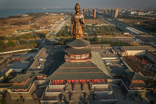 Aerial view of Huayi Brother Film World featuring a massive bronze Guanyin statue standing above traditional Chinese architecture with red curtains and tiled roofs in Suzhou, Jiangsu, China.
