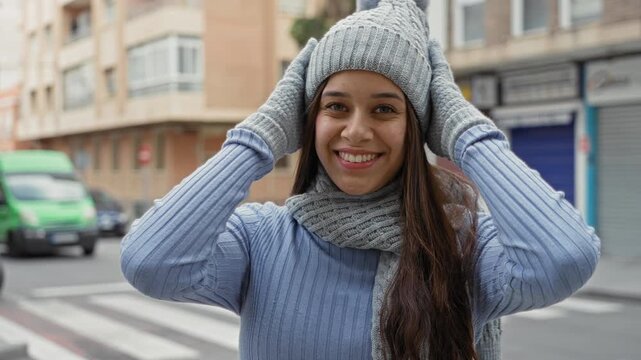 Woman brunette adjusts gray knit hat and gloves, holding hat at temples and smiling on a street crosswalk beside a parked green van and shuttered storefront; winter warmth joy.