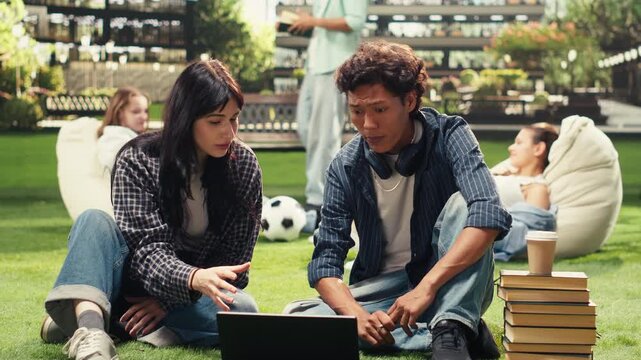 Cheerful students sit on the grass and share ideas in a project discussion, learning together with classmates and friends in an outdoor collaborative study session on a modern university campus.