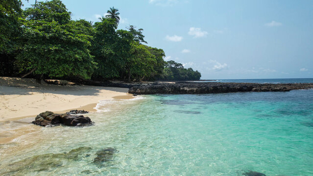 The tranquil shoreline of Praia Caf&eacute; on Ilh&eacute;u das Rolas features transparent turquoise waters, white sand, and volcanic reefs surrounded by dense tropical greenery.S&atilde;o Tom&eacute;,Africa