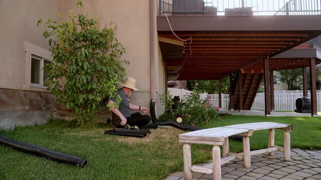 A homeowner sits on the lawn and assembles a black flexible downspout extension beside a residential foundation. The scene shows the drainage components, landscaping, and existing gutter downspout.