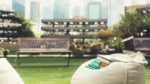 Stacks of books rest on a beanbag on a sunny campus lawn, creating an empty outdoor education scene with blurred modern buildings behind and research tools arranged beside the study materials.