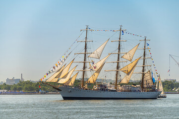 Tall ship USCGC Eagle sails through New York Harbor. Three masted barque displays signal flags and cream sails over blue water with trees and port cranes behind. © white78