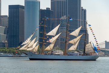 USCGC Eagle passes Lower Manhattan skyline during Independence Day. White tall ship carries cream sails and signal flags across harbor water before glass office towers. © white78
