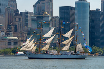 Tall ship crosses New York Harbor before Lower Manhattan towers. USCGC Eagle sails across calm water with cream sails and signal flags against dense city buildings. © white78