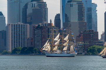 USCGC Eagle sails beneath Lower Manhattan skyscrapers on July 4. Vertical telephoto view places the tall ship below glass towers, brick buildings and waterfront trees. © white78