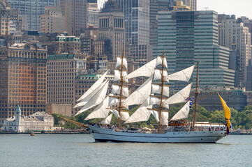 BAE Guayas sails beneath Lower Manhattan towers on July 4. Vertical telephoto view shows the Ecuadorian barque with white sails against historic and glass buildings. © white78