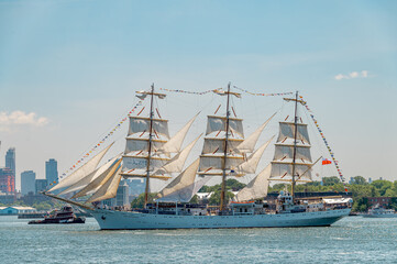 Dar Mlodziezy sails through New York Harbor during Independence Day. Polish training ship carries white sails and signal flags across harbor water with trees and towers behind. © white78