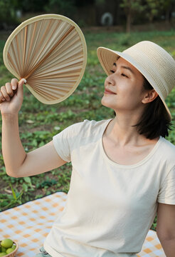 Asian woman in a straw hat fanning herself with a traditional hand fan during a summer picnic outdoors, She sits on a checkered blanket in a green park, enjoying the warm weather and fresh air