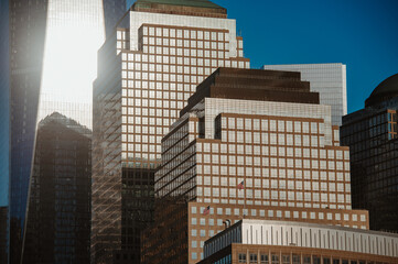 Obraz premium One World Trade Center behind American flags on Independence Day. Warm sunlight reflects across layered office facades beneath a clear blue sky.