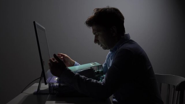 Man working late at night, illuminated by a computer screen, handling documents and ring binders on a desk, representing concepts of overwork and stress