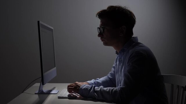 Man in profile wearing glasses working intently on a computer in a dimly lit office, showing dedication and focused concentration on programming or design tasks