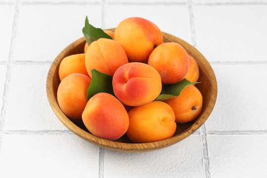 Top view of ripe apricots with green leaves arranged in wooden bowl on on white tile background. Fresh seasonal harvest, summer fruit, healthy eating, dessert, food ingredient