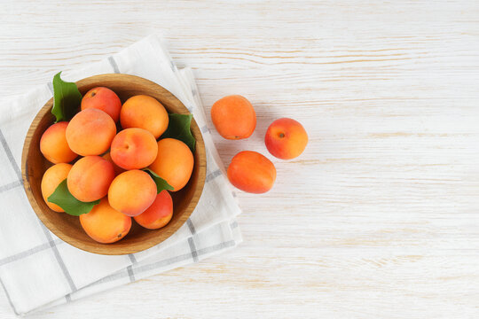 Top view of ripe apricots with green leaves arranged in wooden bowl on on white wooden background with copy space. Fresh seasonal harvest, summer fruit, healthy eating, dessert, food ingredient