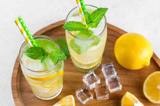 Refreshing homemade lemonade in clear glasses with fresh lemon slices, mint leaves, ice cubes, sparkling water and visible condensation served on wooden tray. Summer citrus beverage, drink concept.