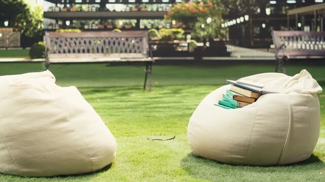 Close-up shows books and research tools on a soft beanbag at a sunny outdoor campus, creating an empty academic study setup with learning materials, literature books and a notebook.
