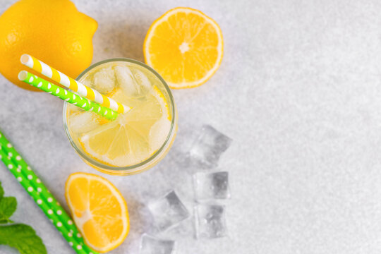 Top view of refreshing cold icy beverage, homemade lemonade with fresh lemon slices, mint leaves, ice cubes, paper straws wtih soda water and condensation in glass on gray background with copy space