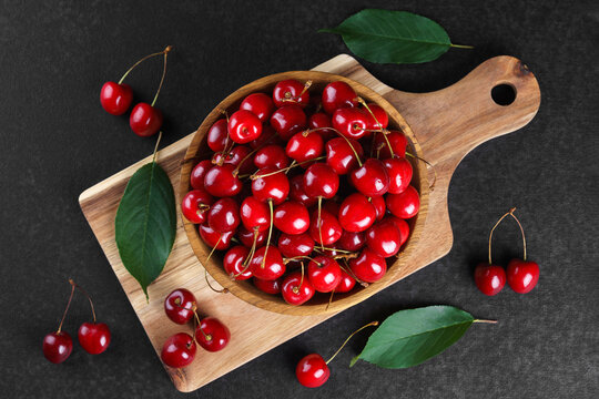 Fresh ripe sweet cherries in wooden bowl on rustic cutting board with green leaves and linen cloth on black textured background. Healthy summer fruit, red berries, fresh seasonal harvest, healthy food
