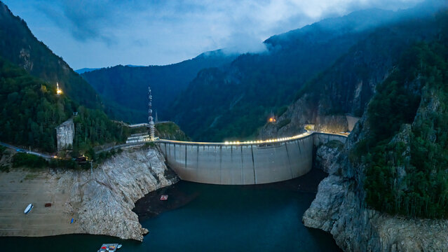Aerial view of the illuminated Vidraru Dam arch structure spanning the reservoir surrounded by steep forested mountains at dusk in Arefu, Arges, Romania.