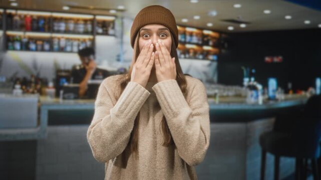 Woman covering mouth with hands at a busy cafe counter with barista and bottles, wearing beanie and cozy sweater; surprise warmth.