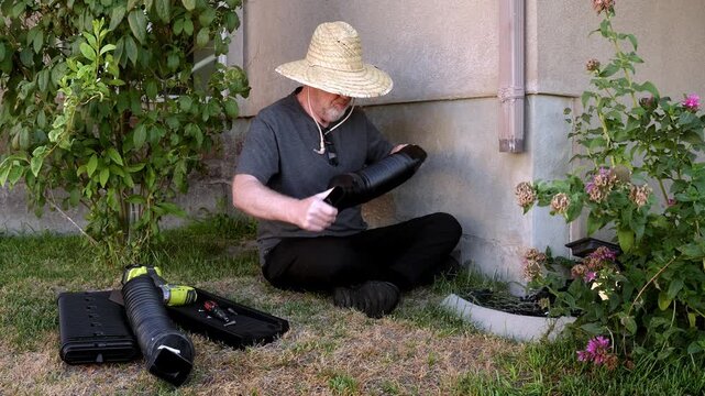 A homeowner sits and assembles a black flexible downspout extension beside a residential foundation. The scene shows the drainage components, landscaping, and existing gutter downspout.
