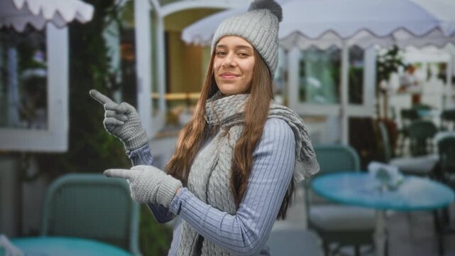 Woman points finger with gloved hand at street cafe table, wearing gray knit hat, scarf and ribbed sweater while smiling; cozy welcome.