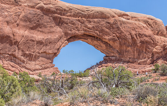 View of North Window Arch with red sandstone formations, desert shrubs, and people visible under the arch in Arches National Park, Grand County, Utah, United States.