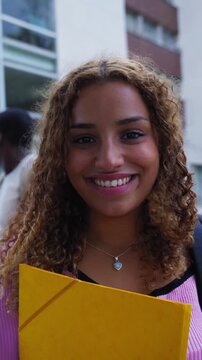 Vertical. Beautiful young african american woman with afro hair smiling and looking at the camera, standing in front of the university building with other students in the background on the stairs